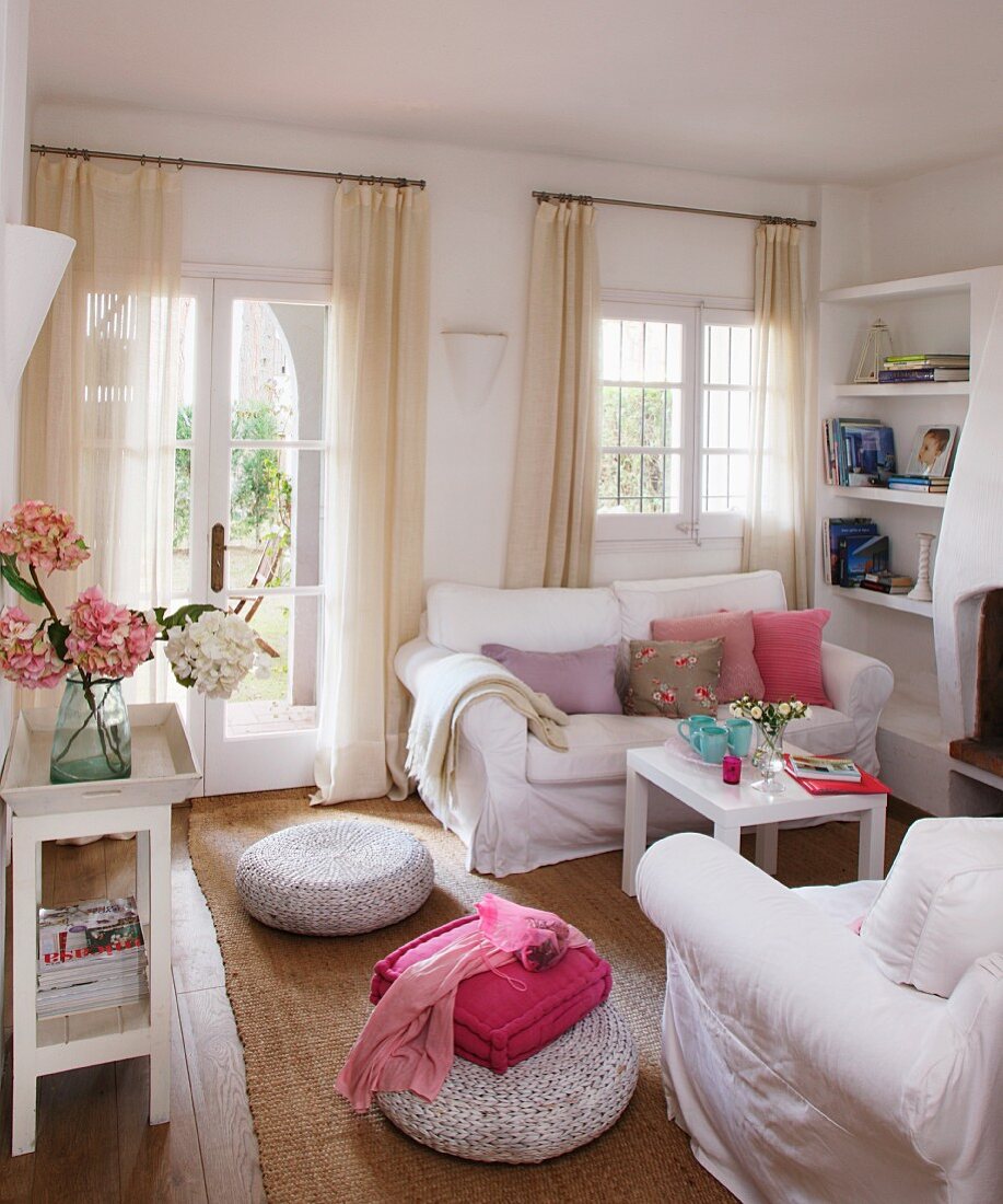 weiße Sofagarnitur und Bodenkissen auf Sisalteppich in Wohnzimmer mit luftigen Vorhängen an Sprossen Terrassentür und Fenster