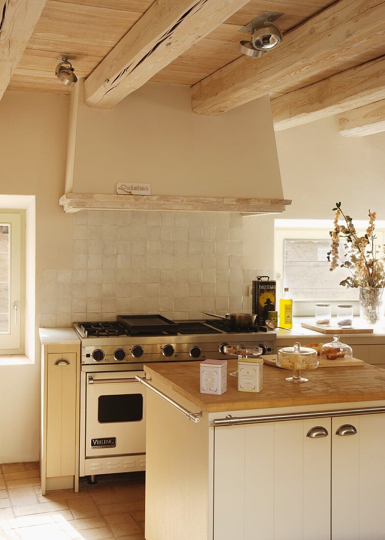 Kitchen island with wooden worksurface in front of cooker with gas hob under extractor hood on wood-beamed ceiling in rustic setting