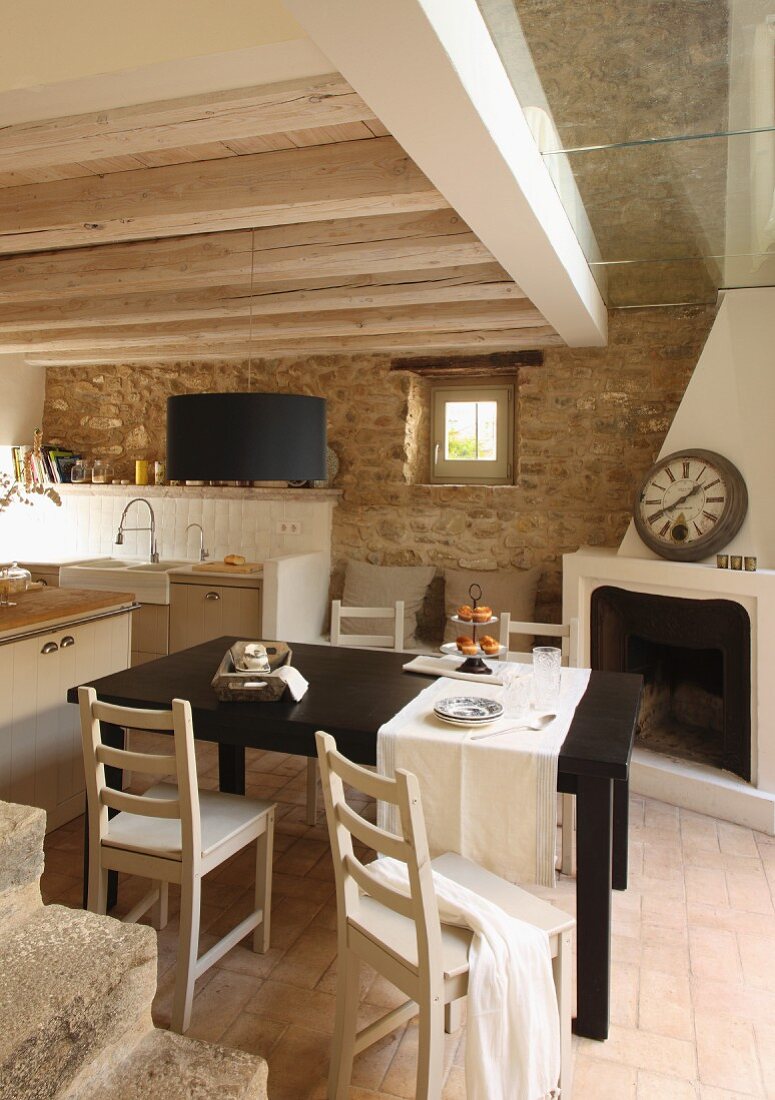Black dining table and simple wooden chairs in front of open, corner fireplace in rustic kitchen with stone wall