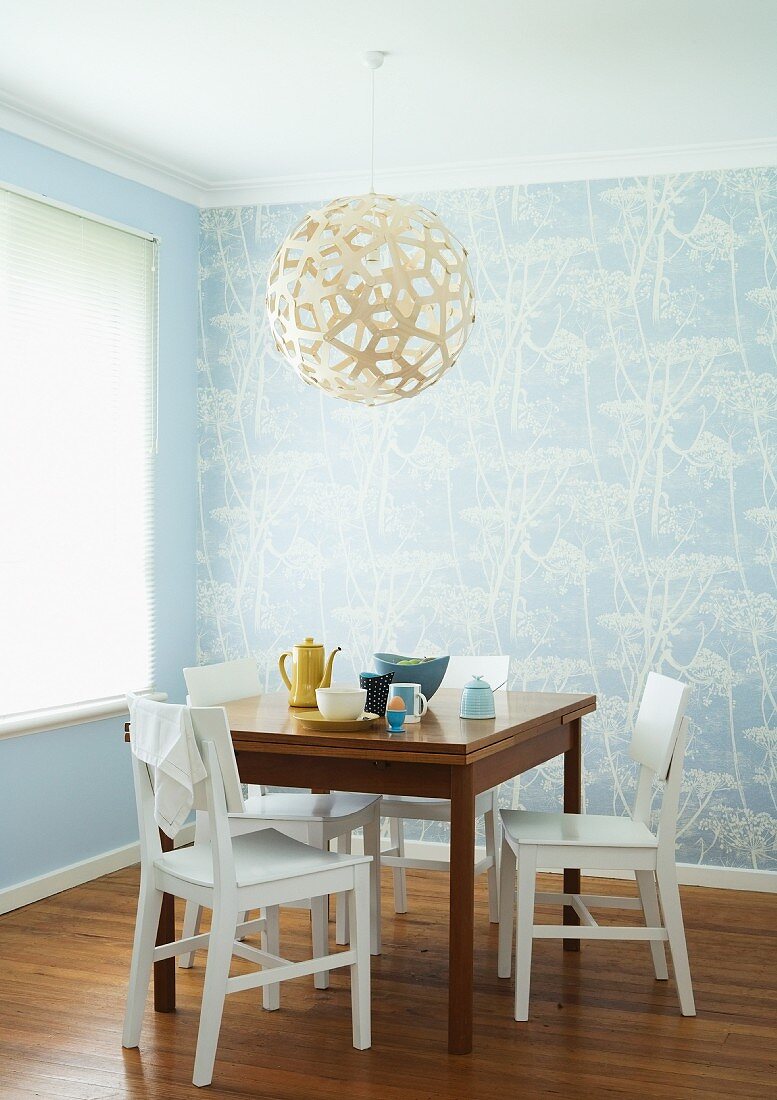 Frühstückstisch und weiße Holzstühle unter kugelförmigem Lampenschirm aus Geflecht in Zimmerecke mit hellblauer Tapete und weißem Blumenmuster an Wand
