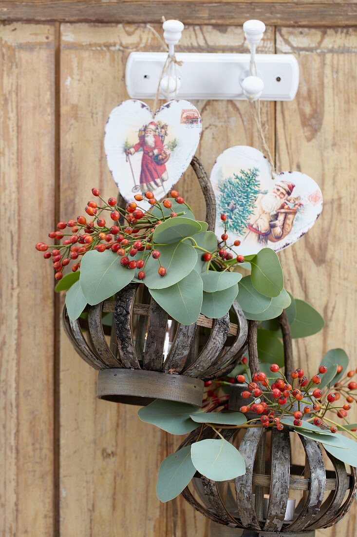 Twigs of rosehips and leaves in baskets and hearts with Christmas motifs hanging from hooks on wooden wall