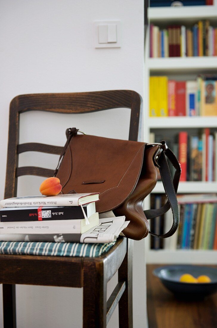 Aprikose auf Bücherstapel und braune Handtasche auf antikem Stuhl an Wand neben offenem Durchgang und Blick auf Bücherregal