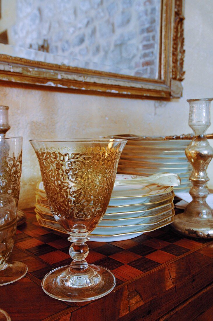 Ornamented wine glasses and gold-rimmed plates stacked on cabinet with chequerboard inlaid top