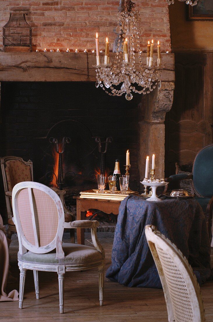 Kronleuchter mit brennenden Kerzen über Rokokostühlchen und Tisch mit drapiertem Tischtuch vor rustikalem Kamin im Wohnzimmer