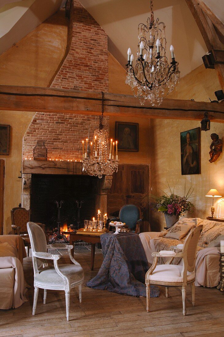 Rustikales Landhaus - Wohnzimmer - Kronleuchter über Sesseln im Rokokostil und schlichter Holztisch mit drapiertem Tischtuch vor offenem Kamin
