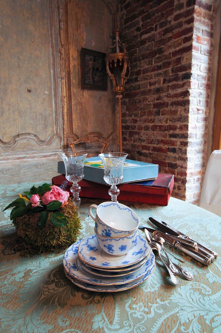 Tasse-n und Untertassenstapel mit weiss blauer Bemalung neben Silberbesteck und Blumengesteck auf Vintage Tischdecke in rustikaler Esszimmerecke