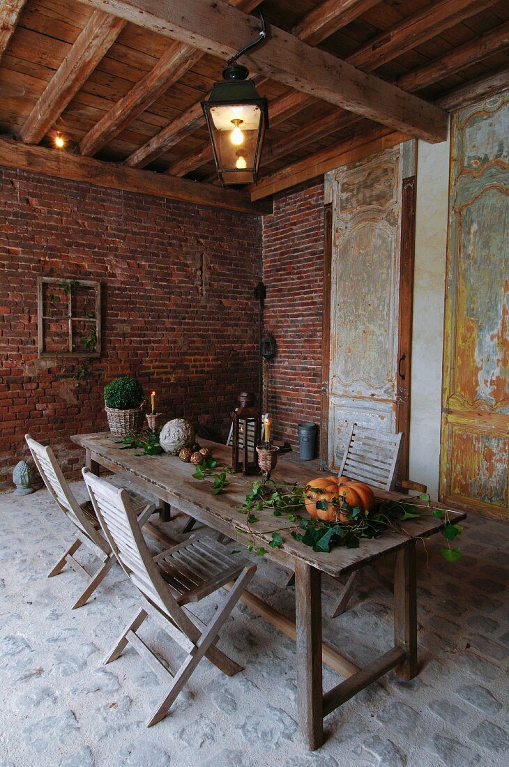 Kürbis und Blumenzweige auf rustikalem Holztisch und Klappstühle in scheunenartigem Raum