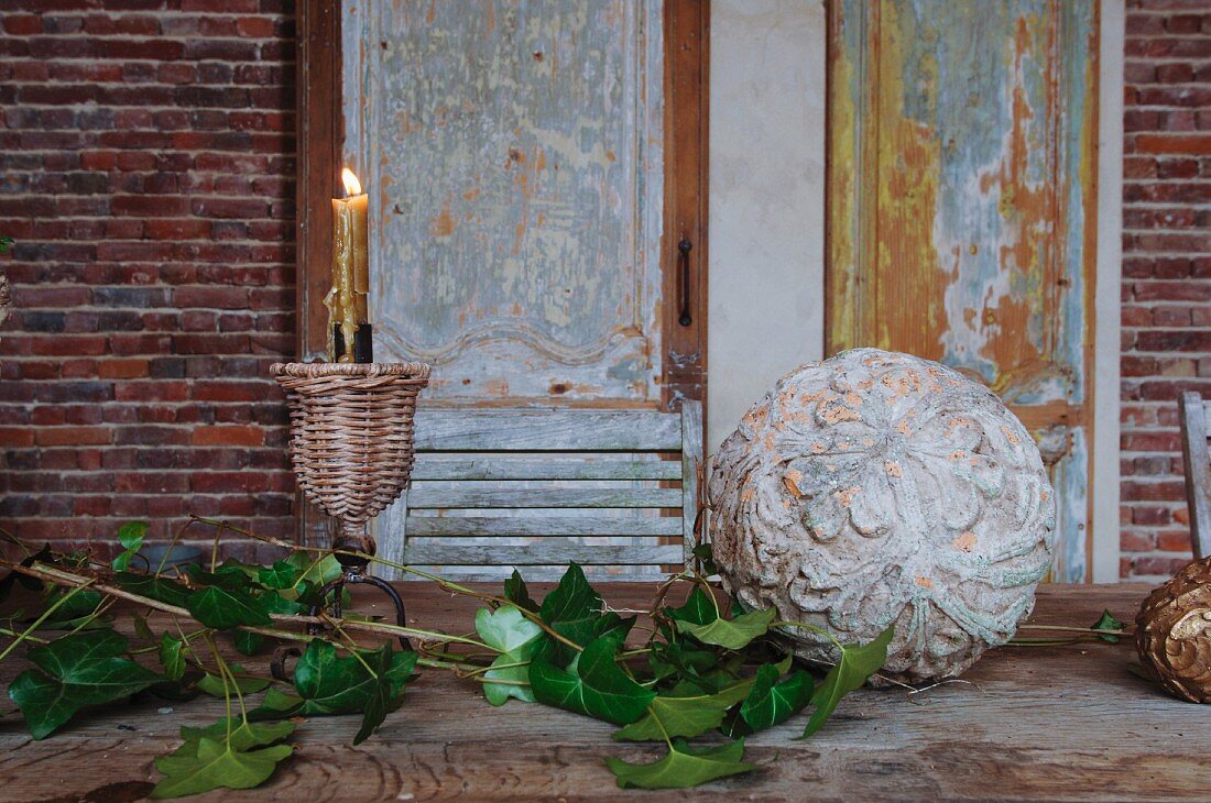 Branches of leaves next to stone ball carved with botanical pattern and lit candle in candlestick on table in front of panelled wooden door with peeling paint