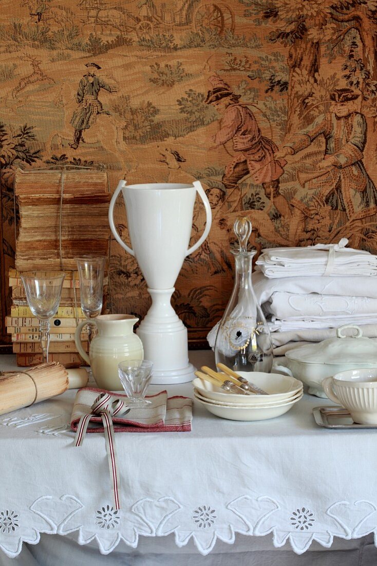Glaswaren und Geschirr, vintage bestickte Tischwäsche, antike Bücher auf einem Tisch vor Wandteppich