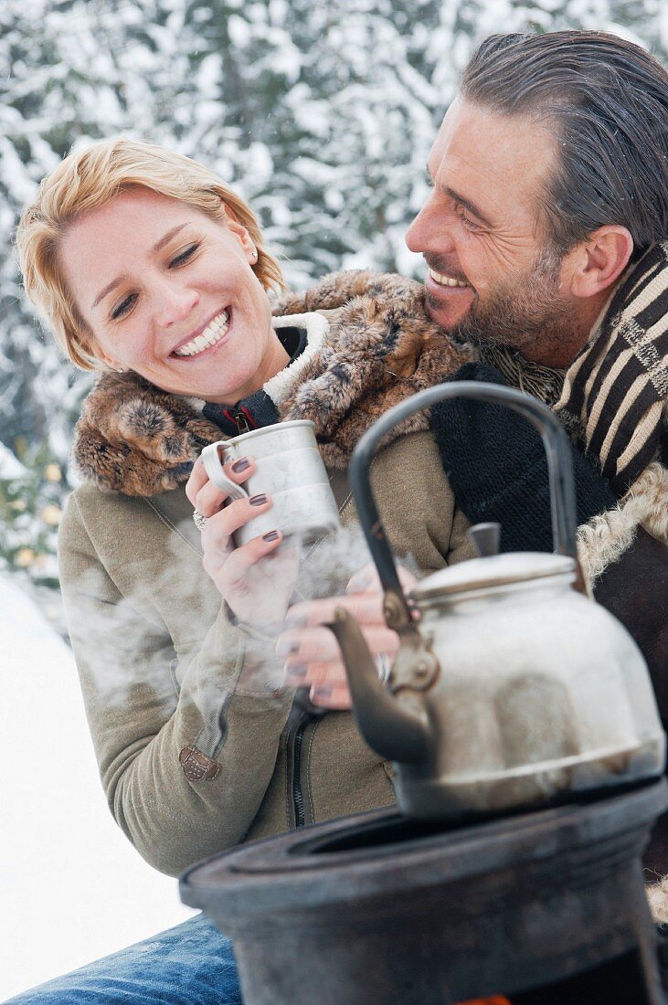 Paar sitzt beim Campingofen im Schnee und trinkt Tee