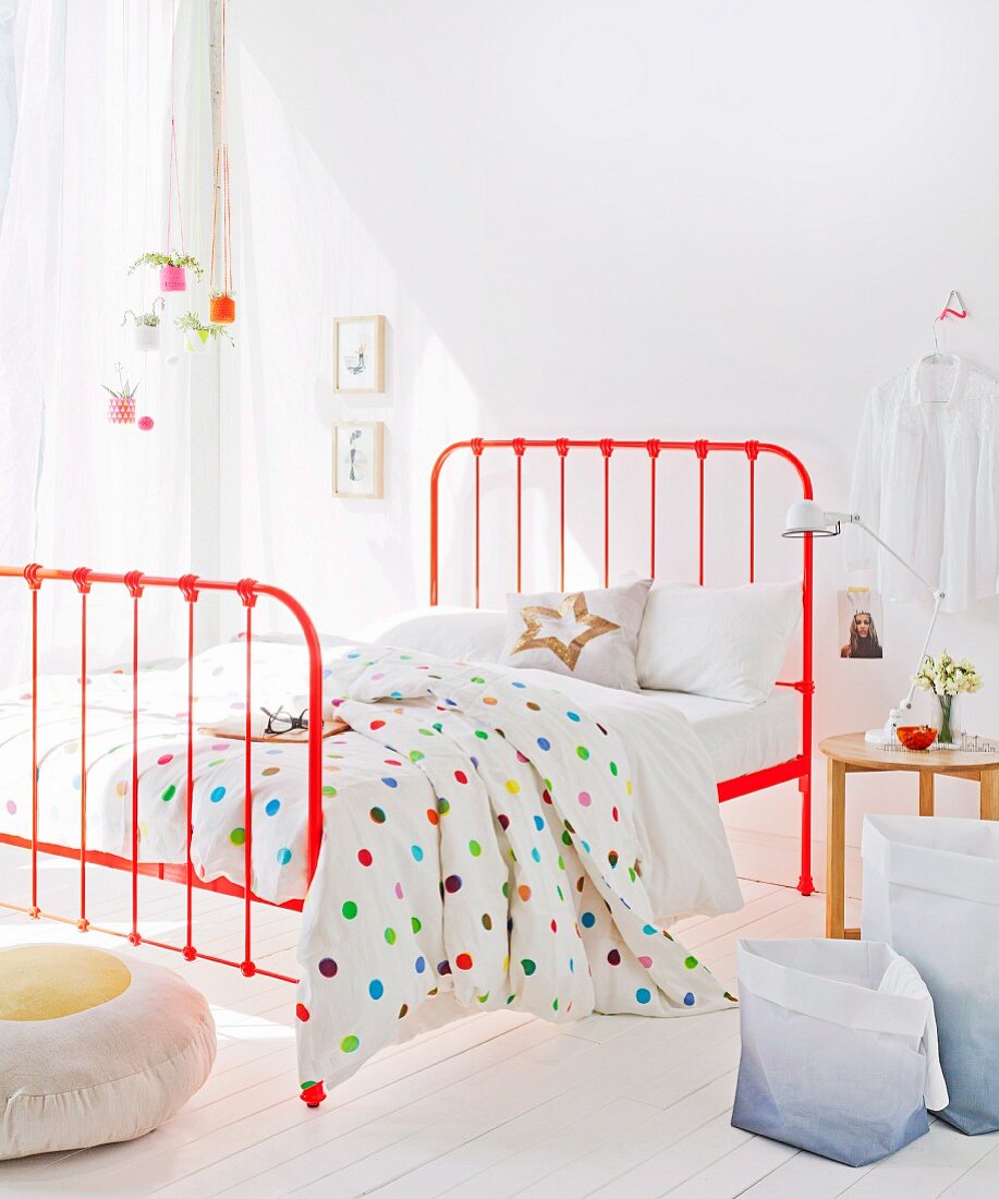 White bed linen with multicoloured polka-dots on orange metal bed in bedroom flooded with light