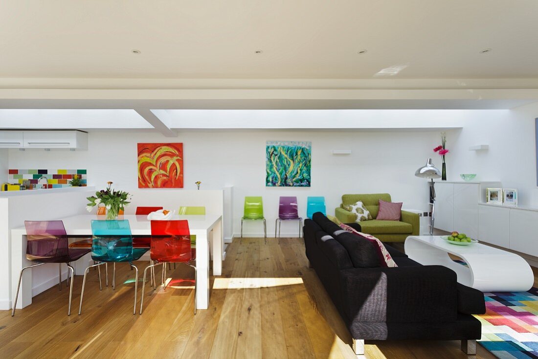 Colourful plastic chairs at white dining table next to lounge area with black couch in open-plan living space