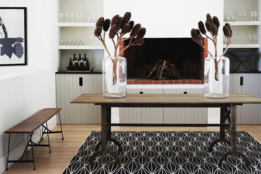 Branches of cones in glass vases on rustic wooden table in front of open fireplace in modern interior