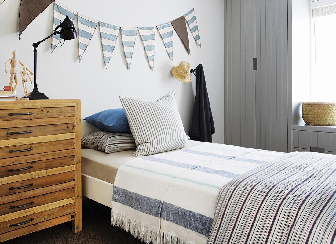 Holzkommode neben Bett mit gestreifter Bettwäsche vor Wand mit Wimpelgirlande