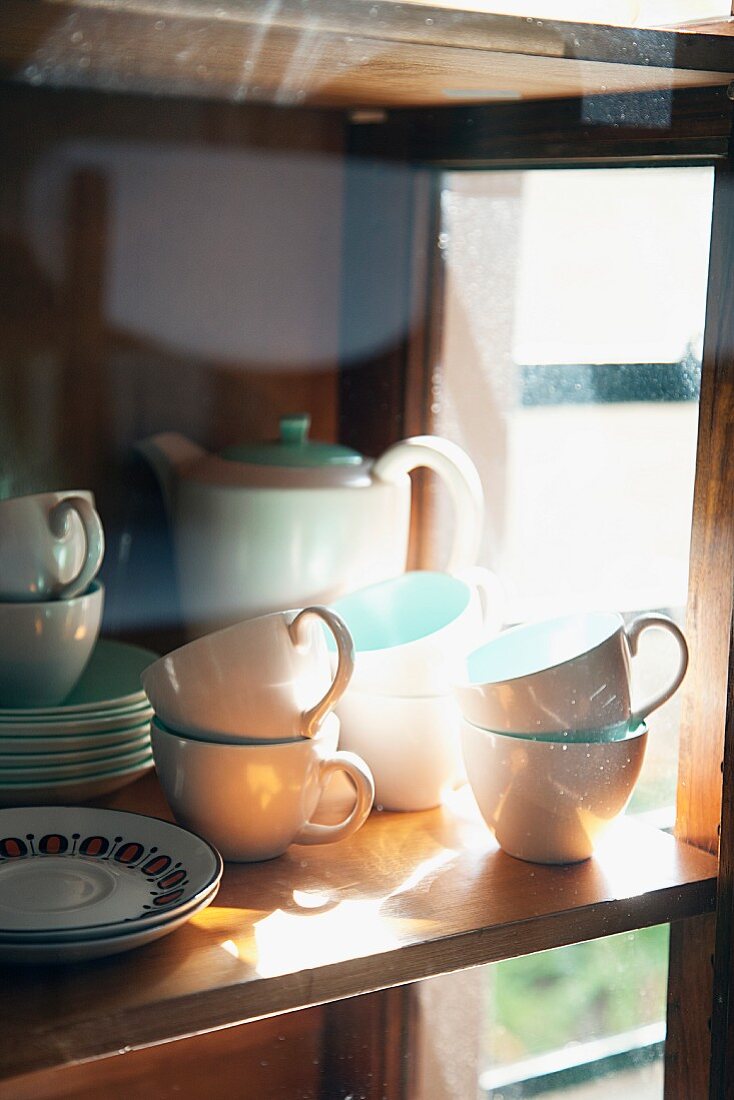 Kaffeegeschirr in Weiß und Pastelltürkis hinter spiegelndem Glas in einfachem Holzregal