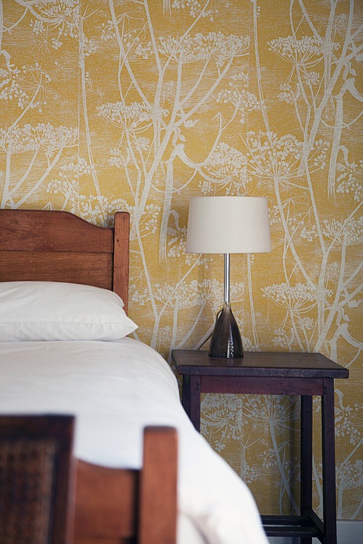 Wooden bed and lamp on bedside table against floral wallpaper