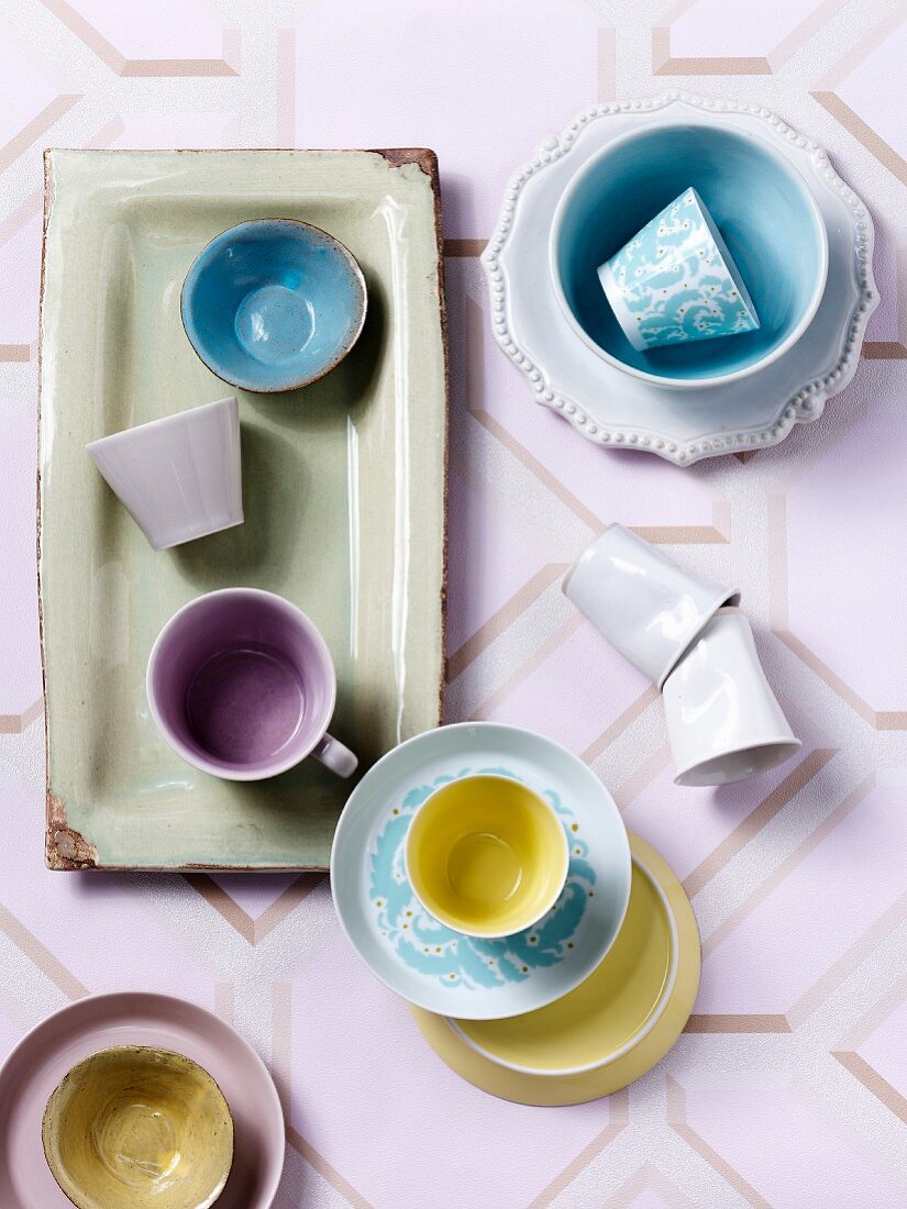 Blick auf pastellfarbene Tassen, teilweise auf Schalen und Untertassen auf gemusterter Tischdecke im Retrostil