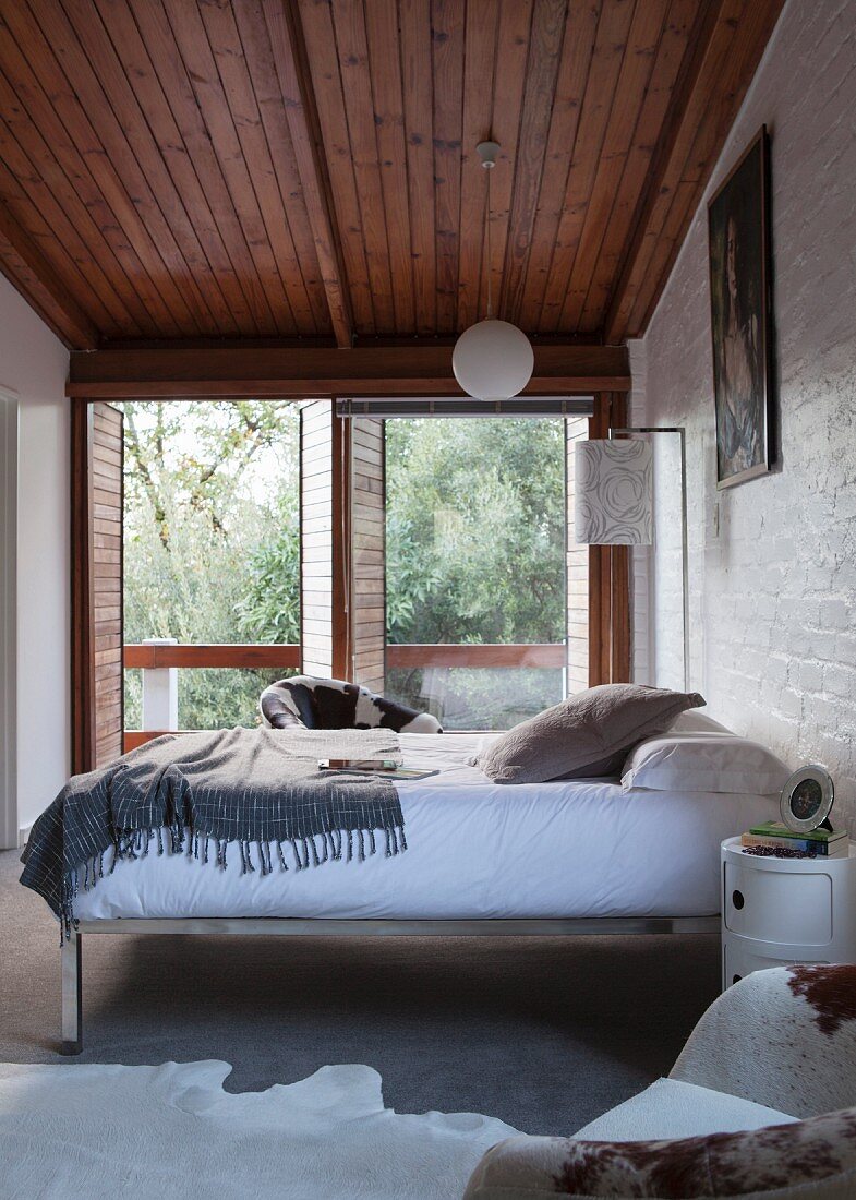 Bed with chrome metal frame against wall in front of terrace doors in plain bedroom with wooden ceiling