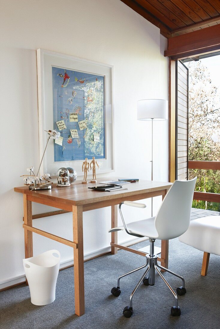 Moderner Büro Drehstuhl an Holztisch vor Wand mit aufgehängtem Bild neben Stehleuchte und offene Balkontür