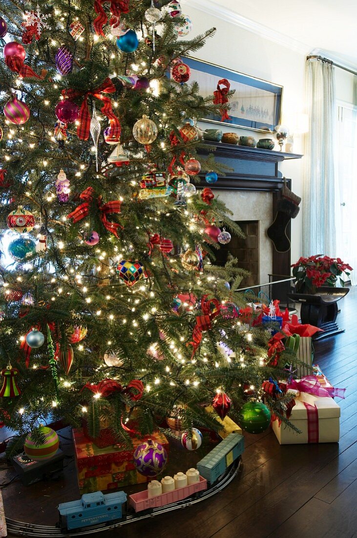 Spielzeugeisenbahn unter leuchtendem, bunt geschmücktem Weihnachtsbaum vor traditionellem Kamin