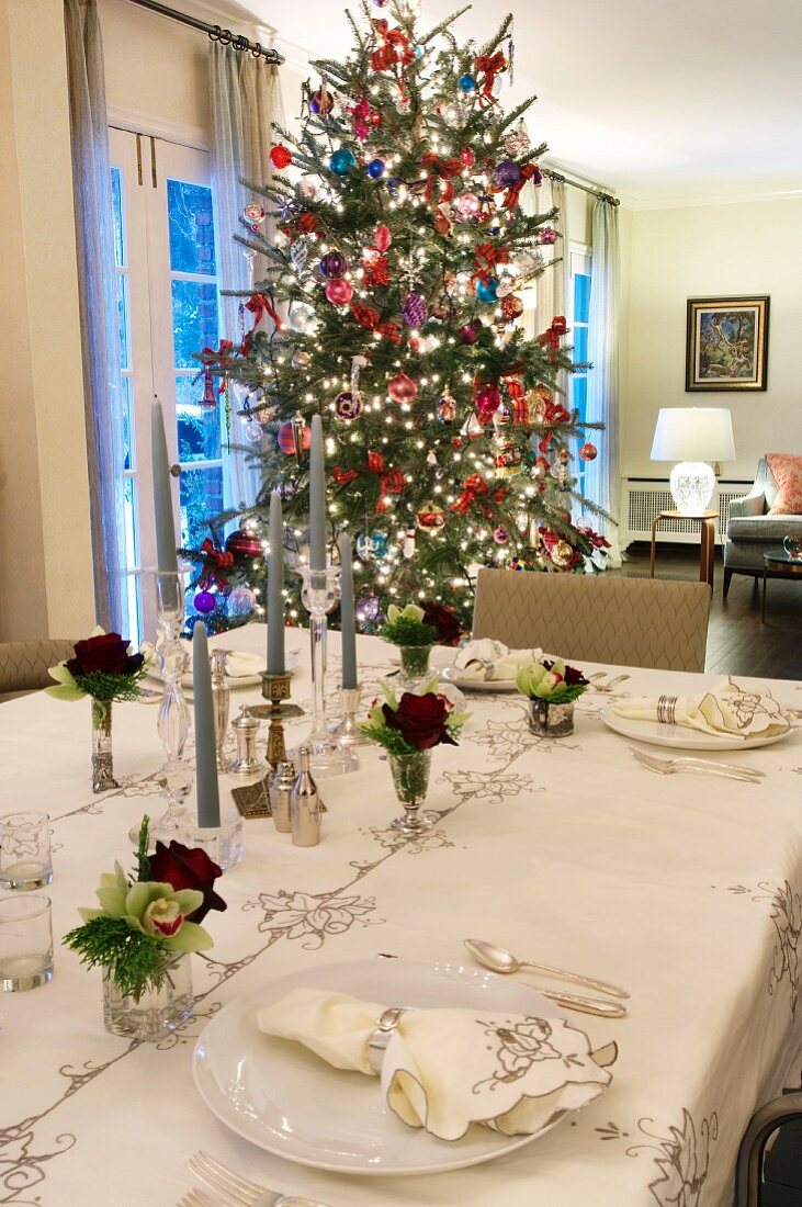 Dezent gemusterte Tischdecke mit Servietten und Rosendeko auf festlicher Tafel mit Weihnachtsbaum im Hintergrund