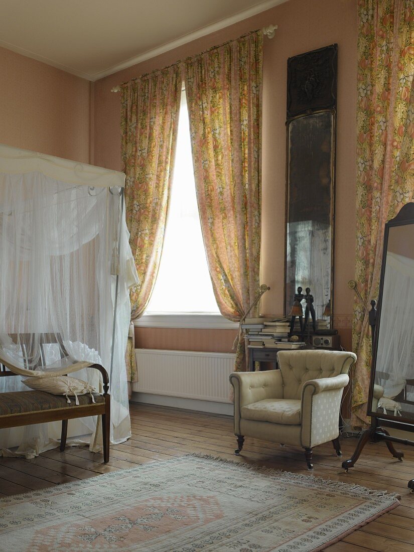 High-ceilinged bedroom with gathered curtains and walls painted in pastel pink and apricot; antique armchair and four-poster bed with white voile canopy