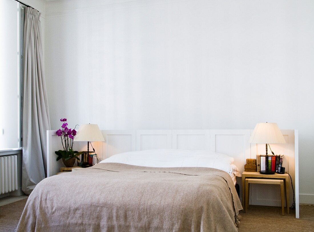 Doppelbett mit hellbrauner Tagesdecke vor halbhoher, weisser Kassettenwand in minimalistischem Schlafzimmer