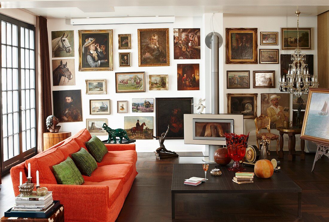 Große Ölbildersammlung in Wohnzimmer mit orangefarbener Couch, Antiquitäten, Kristallleuchter und Kaminofen