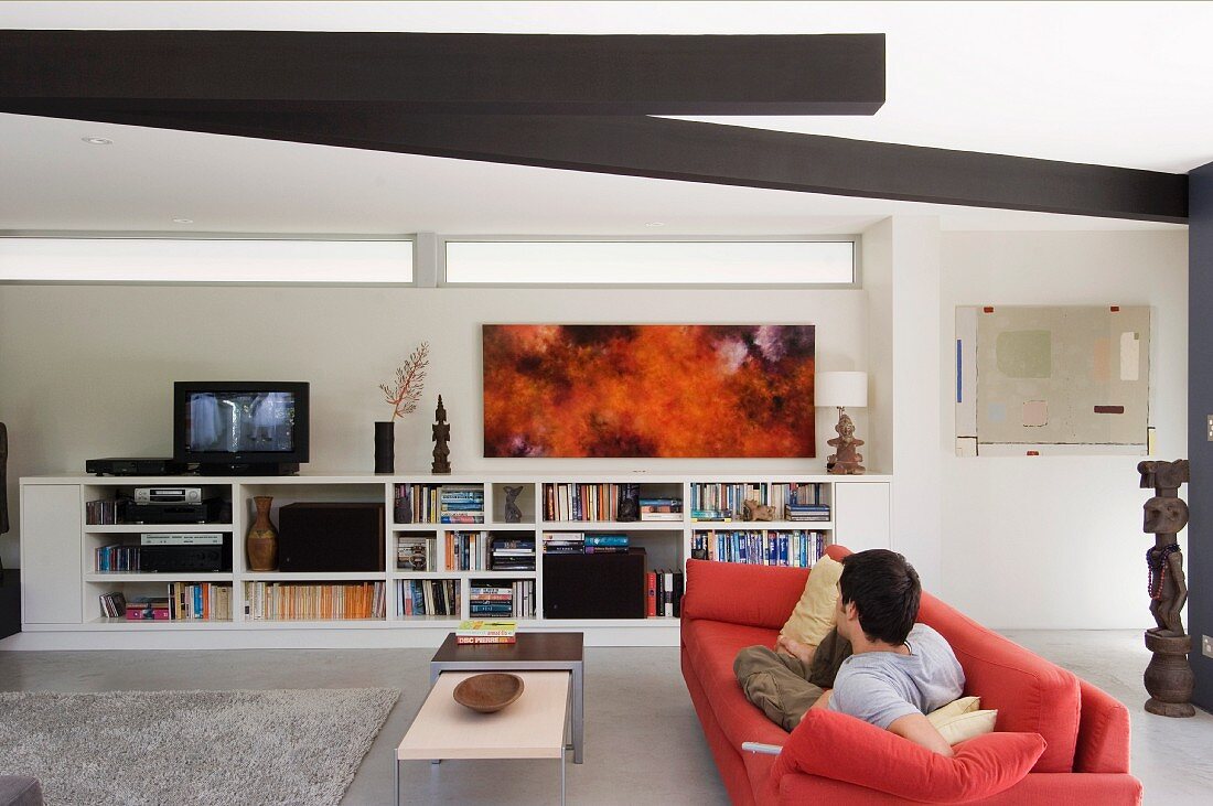 Junger Mann auf rotem Sofa mit Blick auf Fernseher in Medienregal