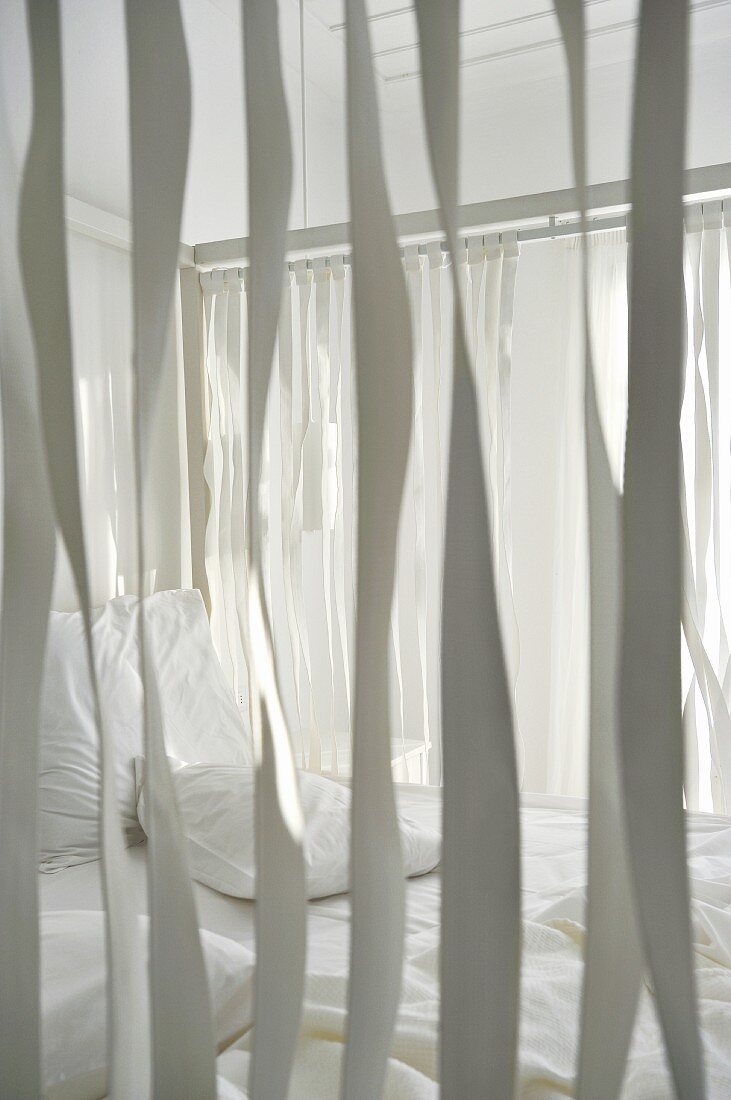 Blick durch aufgehängte Bänder an Holzgestell auf das Himmelbett mit weisser Bettwäsche