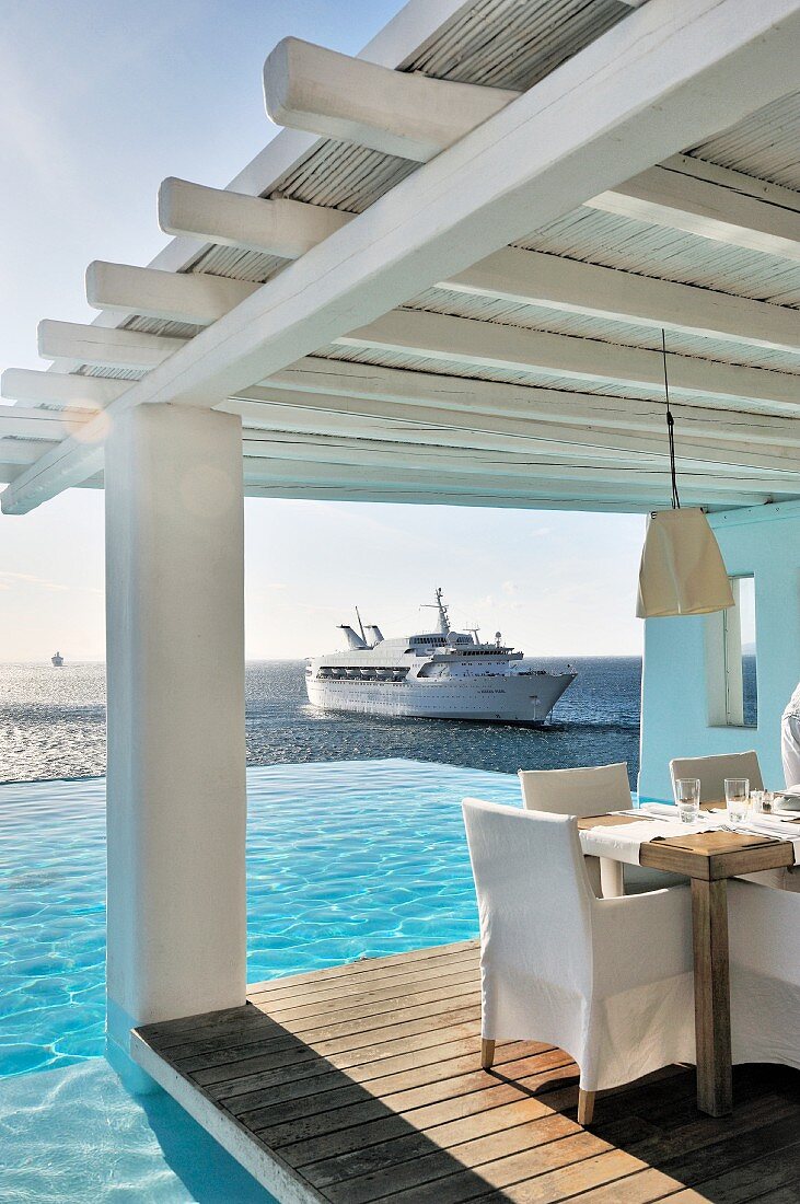 Set table and chairs with white loose covers on roofed wooden terrace next to pool with view of yacht on ocean