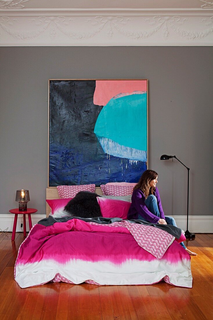 Double bed with pink, batik-style bedspread below large, colourful painting; woman sitting on bed