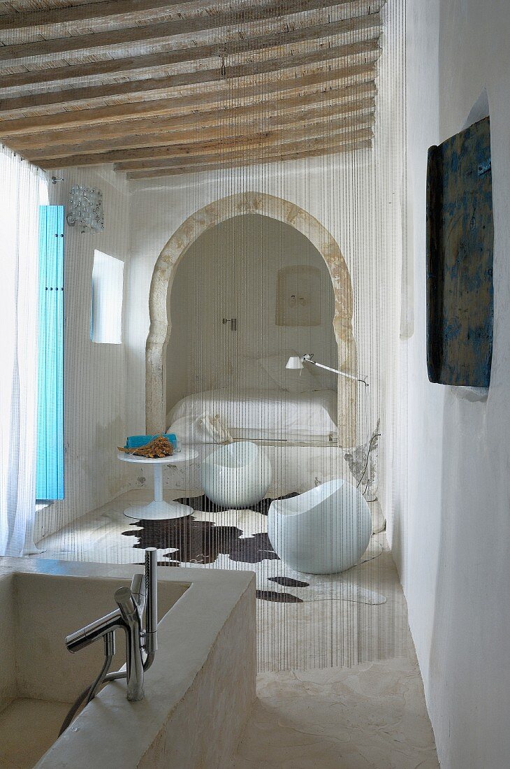 Bad Ensuite mit Natursteinwanne; Schnurvorhang als Abgrenzung zu Sitzplatz mit Designermöbeln und Bett in orientalischem Alkoven
