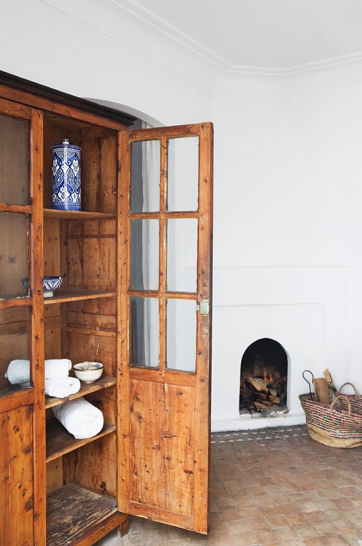 Alter Vitrinenschrank mit offener Tür in Wohnraum mit Terracottaboden und gemauertem offenem Kamin