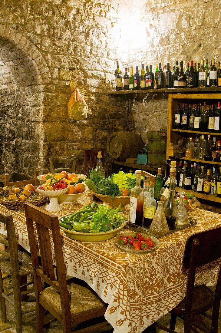 Dishes of vegetables and fruit next to tray of vinegar and oils bottles on table in front of bottles of wine on shelving in rustic dining room