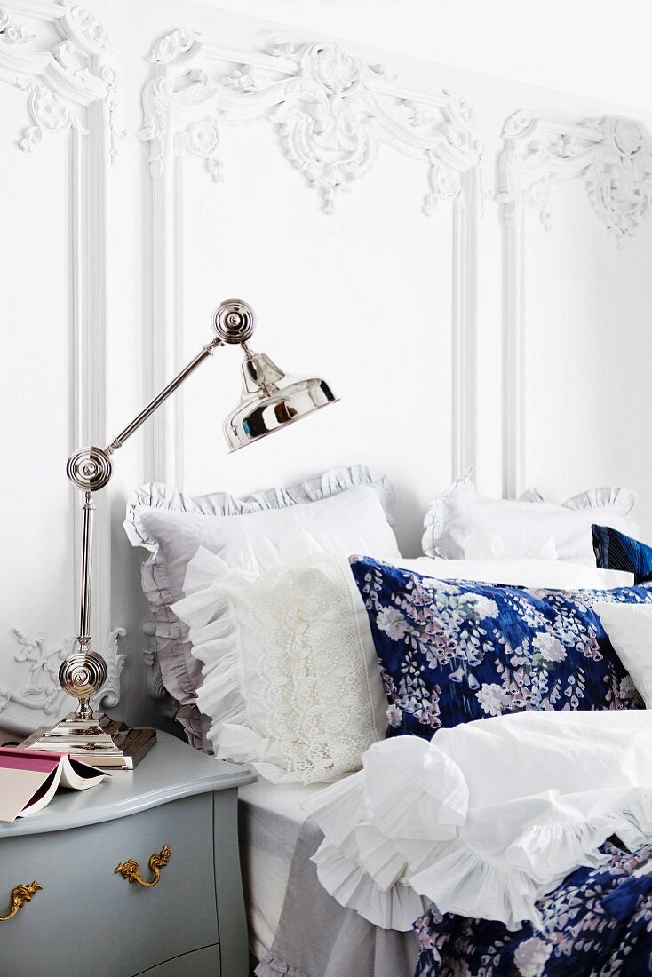 Nostalgisches Doppelbett mit Rüschen- und Spitzenkissen an weisser Wand mit barocker Wanddekoration; auf dem Nachttisch eine verchromte Gelenklampe
