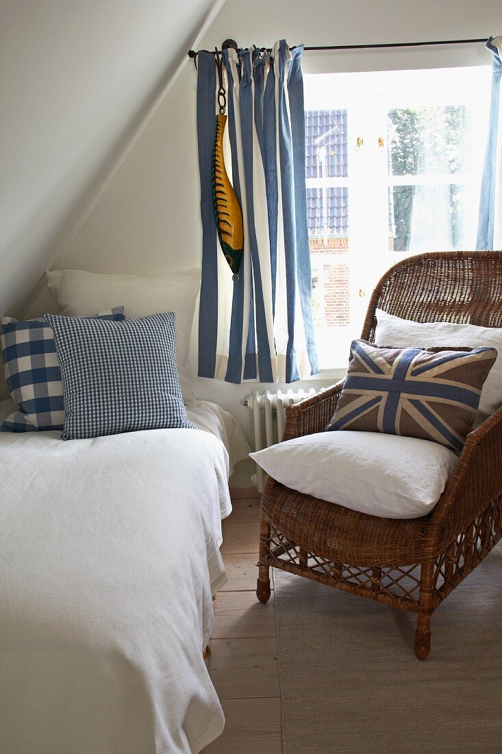 Blue-patterned scatter cushions on comfortable bed and wicker chair under sloping attic ceiling