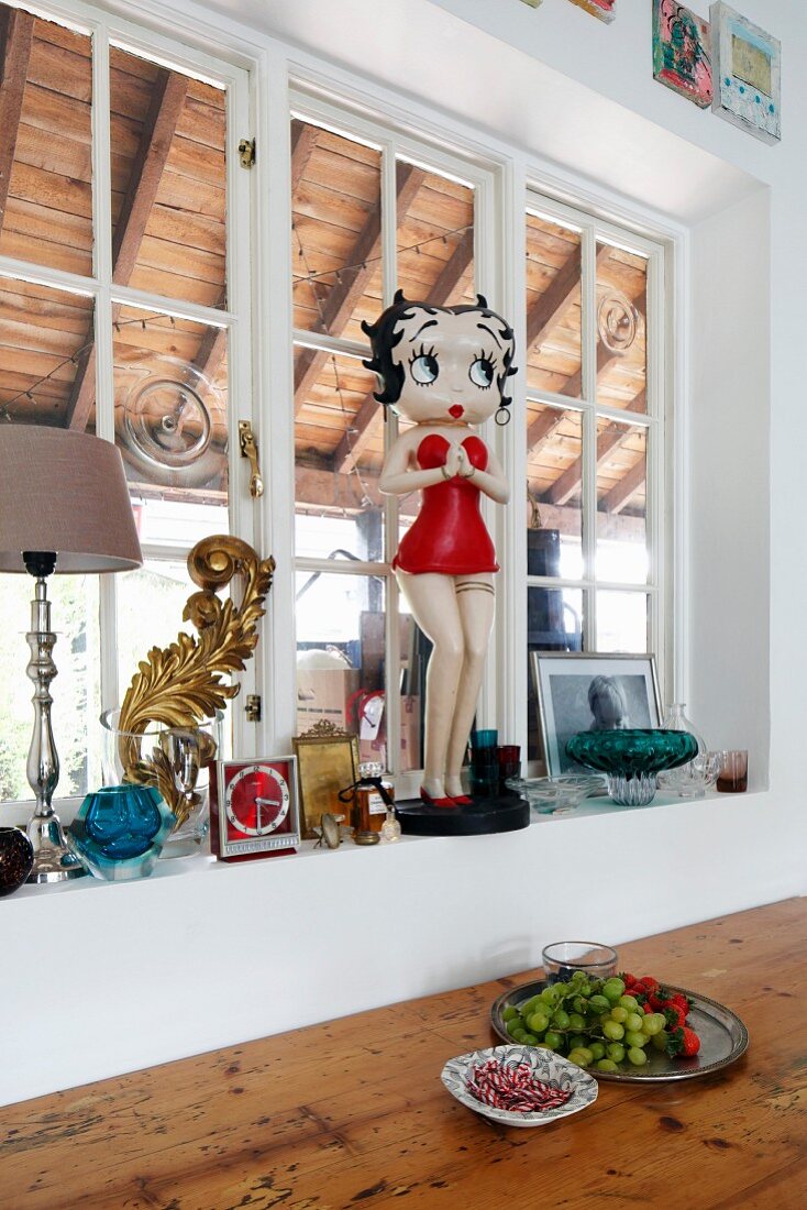 Plastic 'Betty Boop' statue on windowsill of lattice window above platter of fruit on wooden table