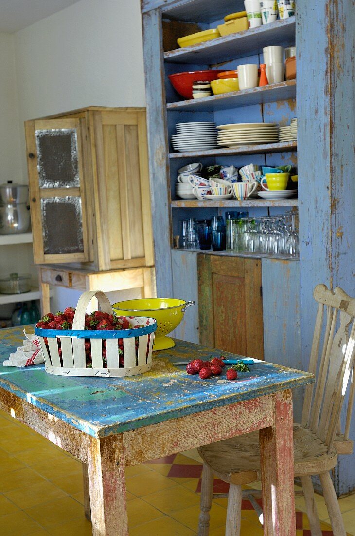 Vintage Küche - Erdbeeren im Korb auf Esstisch mit abblätternder Farbe vor rustikalem Küchenbuffet mit Geschirr