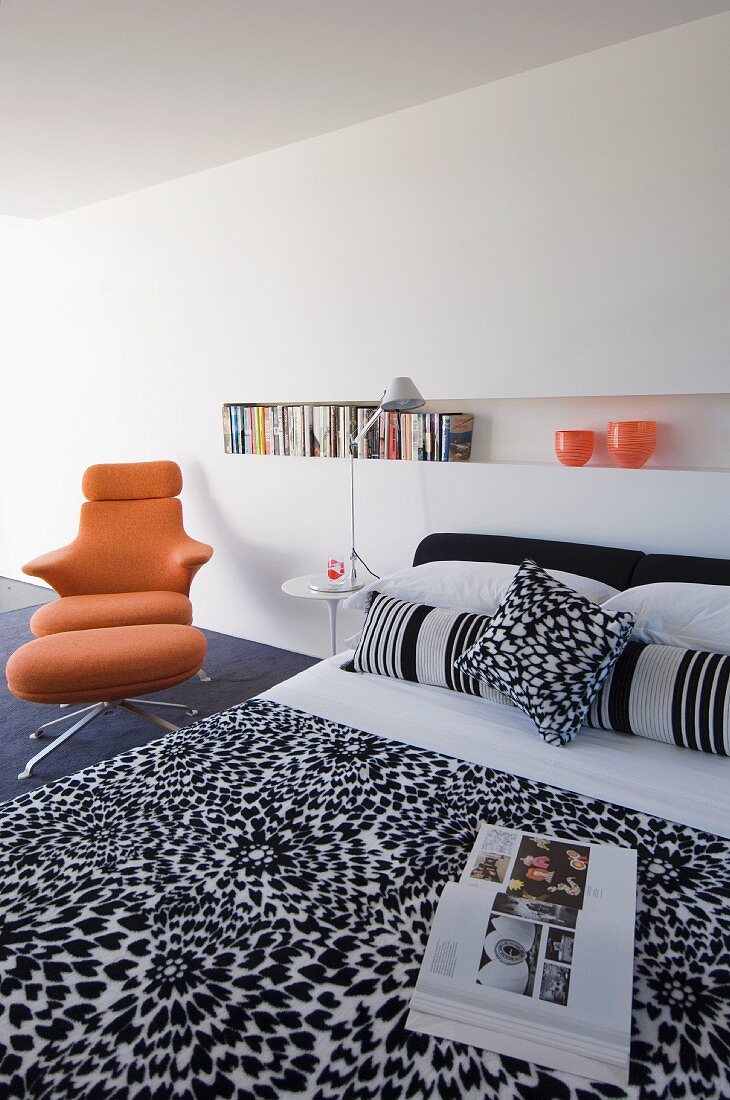 Minimalist bedroom with black and white patterned bedspread, long bookshelf in niche and orange designer swivel chair