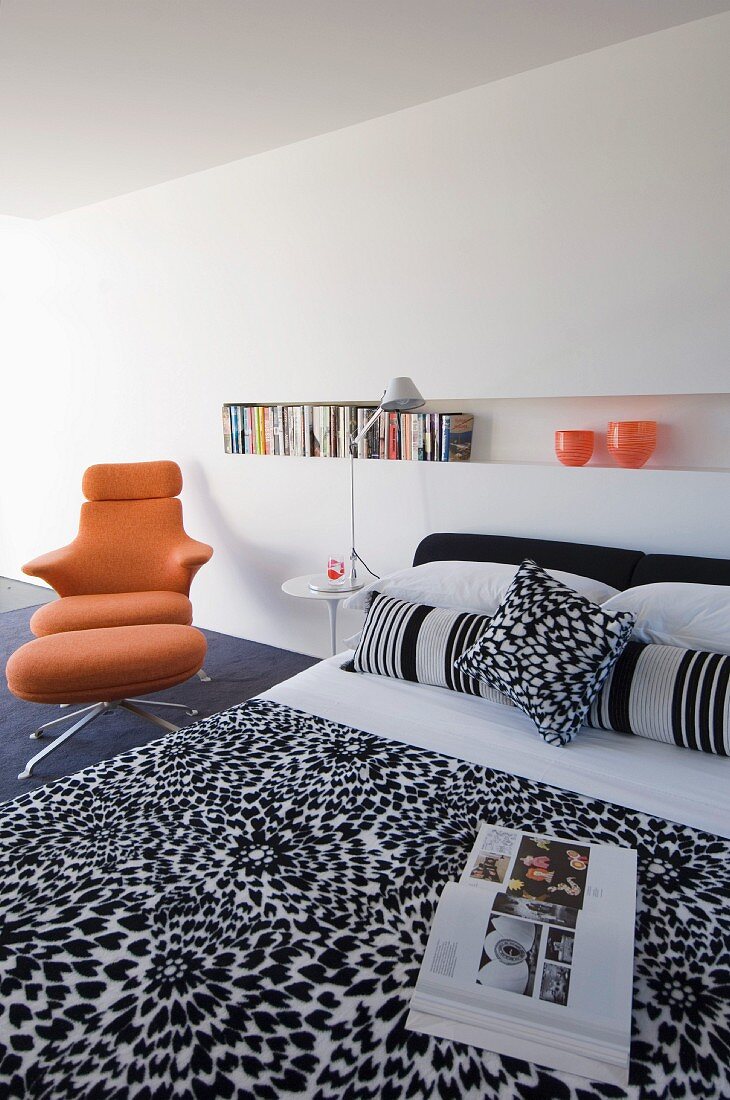 Minimalistisches Schlafzimmer mit schwarz-weiss gemustertem Bettüberwurf, einer Wandnische als Bücherregal und einem orangefarbenen Designerstuhl