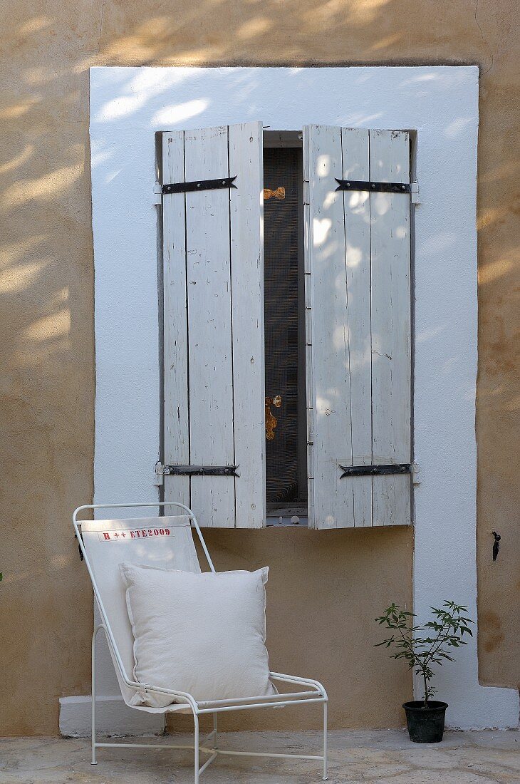 Kissen auf filigranem, weißem Metallstuhl vor Hauswand unter Fenster mit geschlossenen, weissen Holzläden