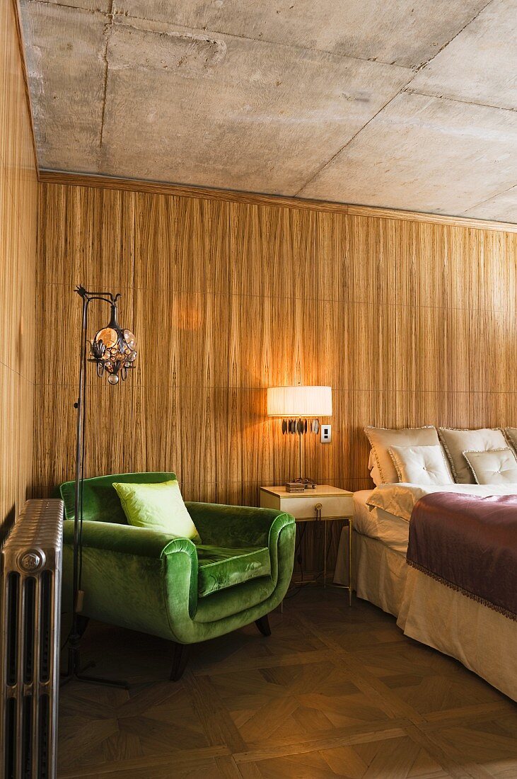 Grüner Sessel und Nachtkästchen neben Bett vor Holzpaneelwand in loftartigem Ambiente