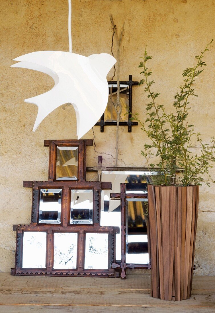 An rustikale Wand gelehnte, originelle Spiegelsammlung; davor eine weiße Hängelampe in der Form eines Vogels und eine hohe Bodenvase aus Holz