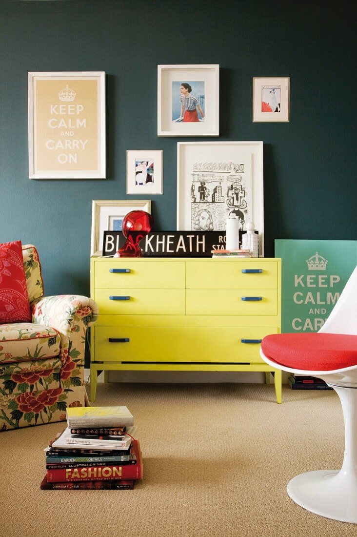 Gelbe Kommode an grauer Wand mit daran hängender Bildersammlung; davor ein Polstersessel mit Blumenmotiv und ein weisser Designer-Drehstuhl mit rotem Sitzkissen