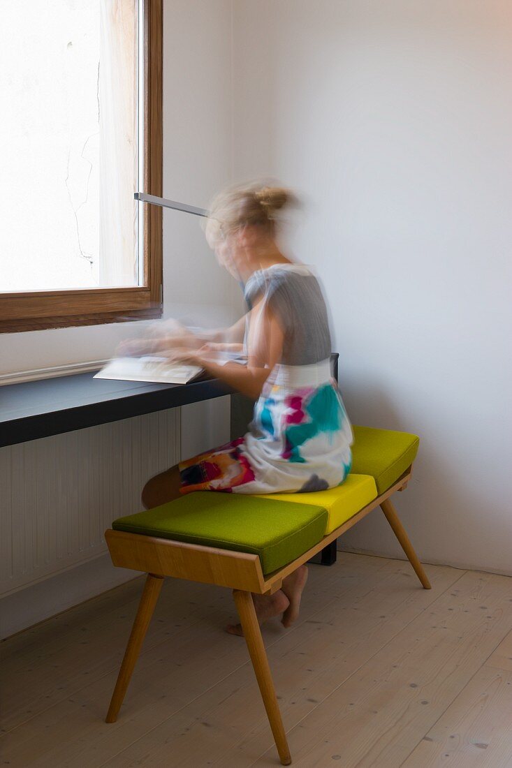 Frau sitzt auf Bank mit grünen Polstern an minimalistischem Tisch vor dem Fenster