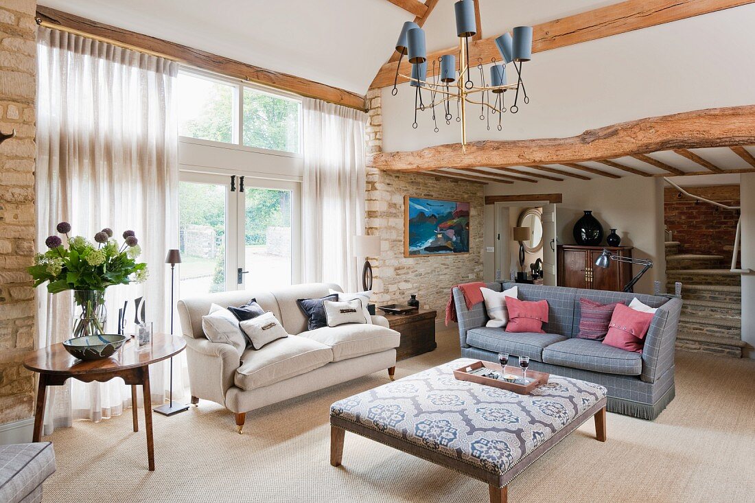 Elegantes Wohnzimmer mit rustikalen Steinwänden und Holzbalken; in der Raummitte zwei Sofas im Vintage-Stil und ein quadratischer Polstertisch