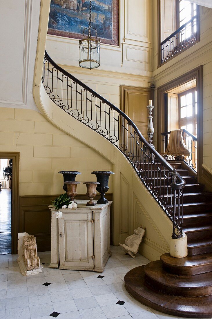 Foot of winding staircase in foyer of stately home with amphorae-style vessels on half-height vintage cabinet