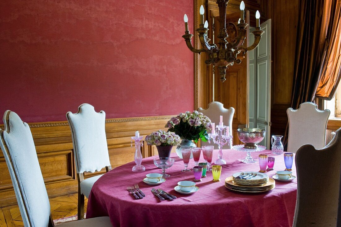 Chairs with white upholstered backrests around table with deep pink tablecloth and places being set in front of wall painted rust red in grand dining room