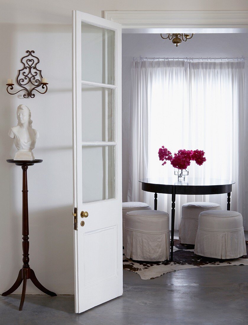 View into white dining room with round table, upholstered stools with white, loose covers; bouquet of bougainvillea on table